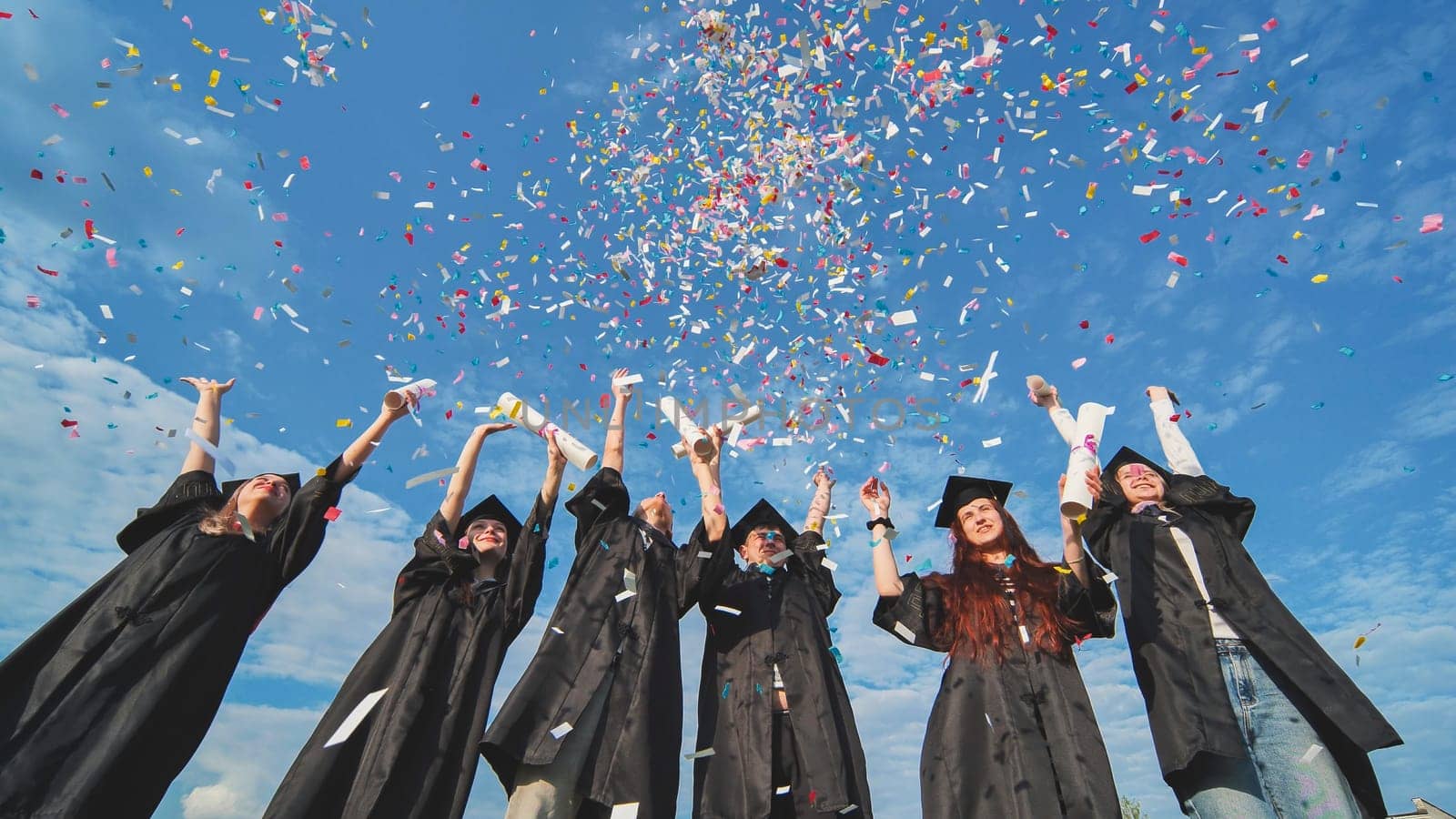 Graduates throw colorful confetti against a blue sky. by DovidPro