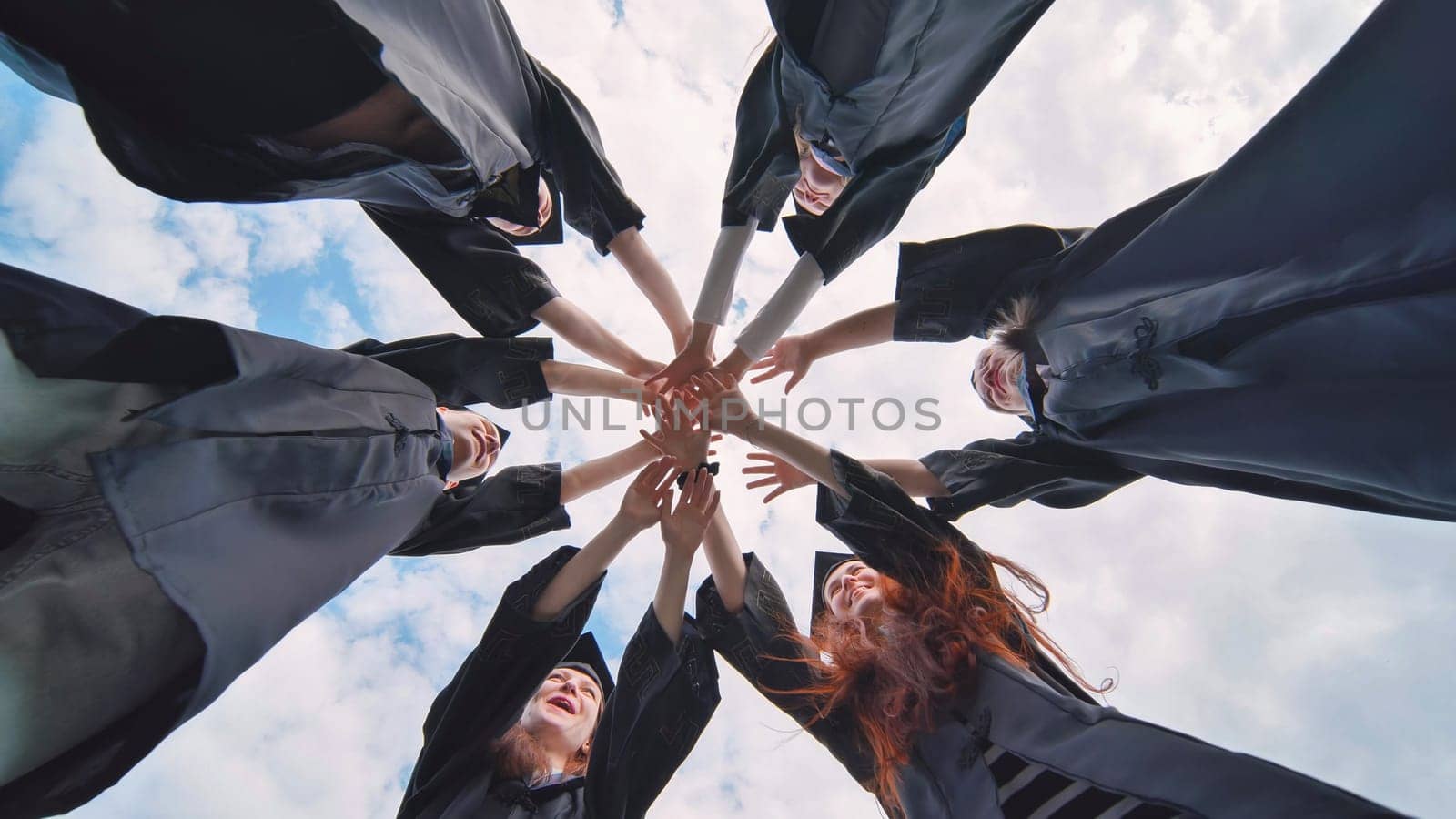 Team of college or university students celebrating graduation. Group of happy successful graduates in academic hats and robes standing in circle and putting their hands together