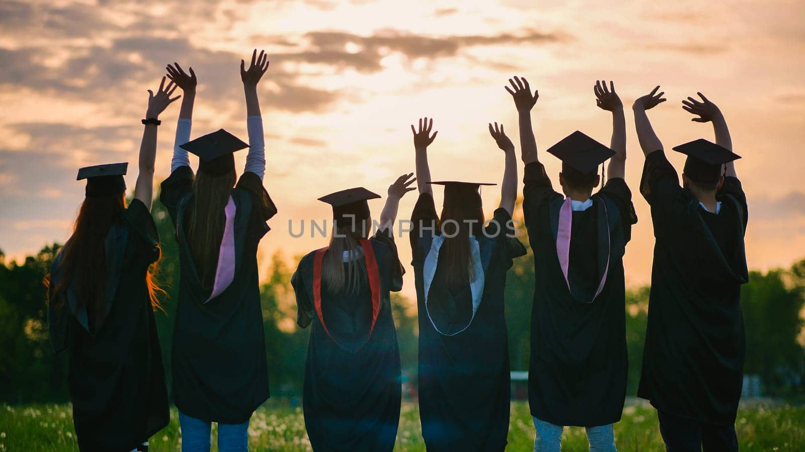 Silhouettes of graduates in black robes waving their arms against the evening sunset