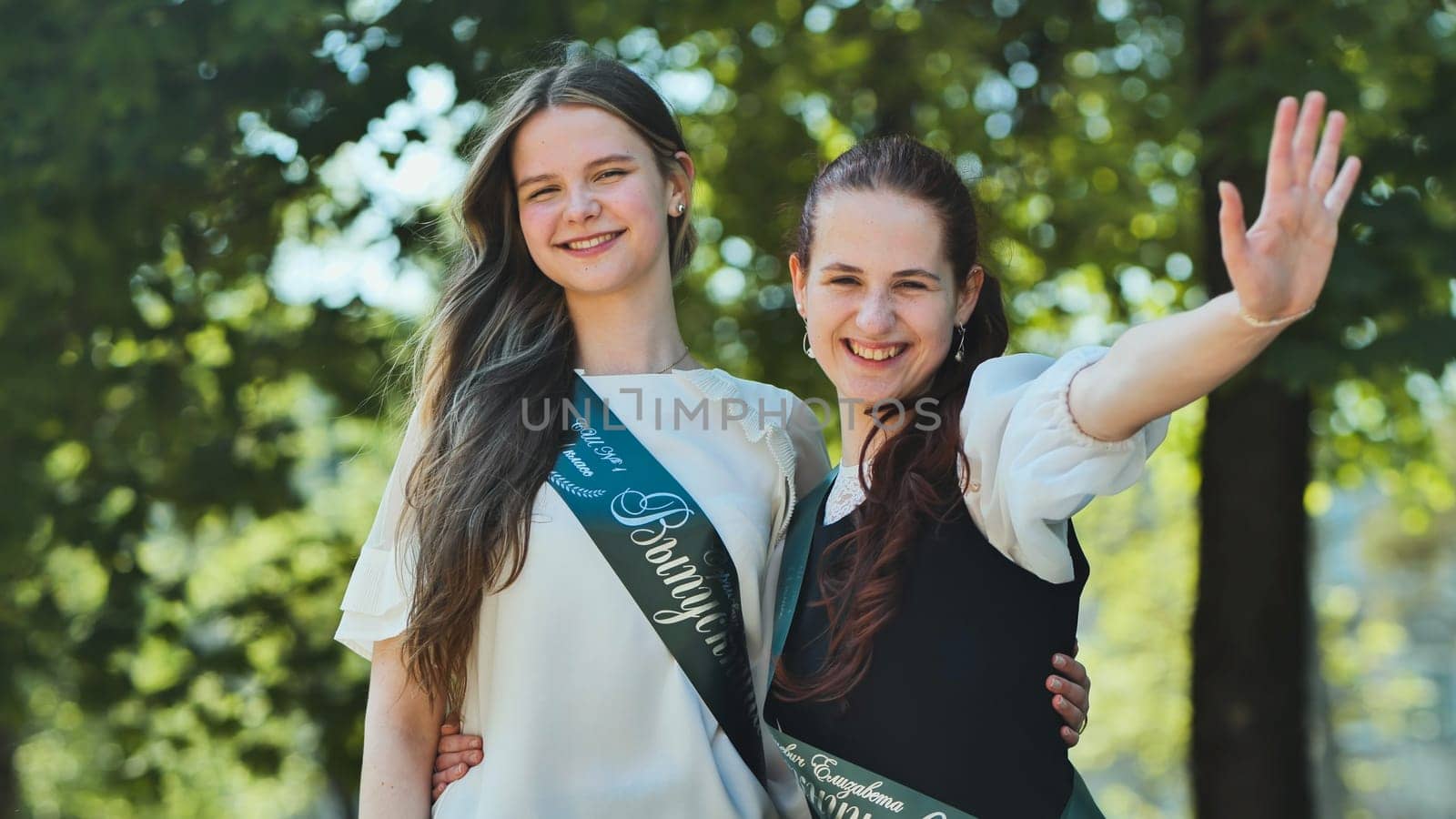 Two Russian schoolgirls graduate posing on a summer day with a ribbon and the name - Graduate 2023 on it. by DovidPro