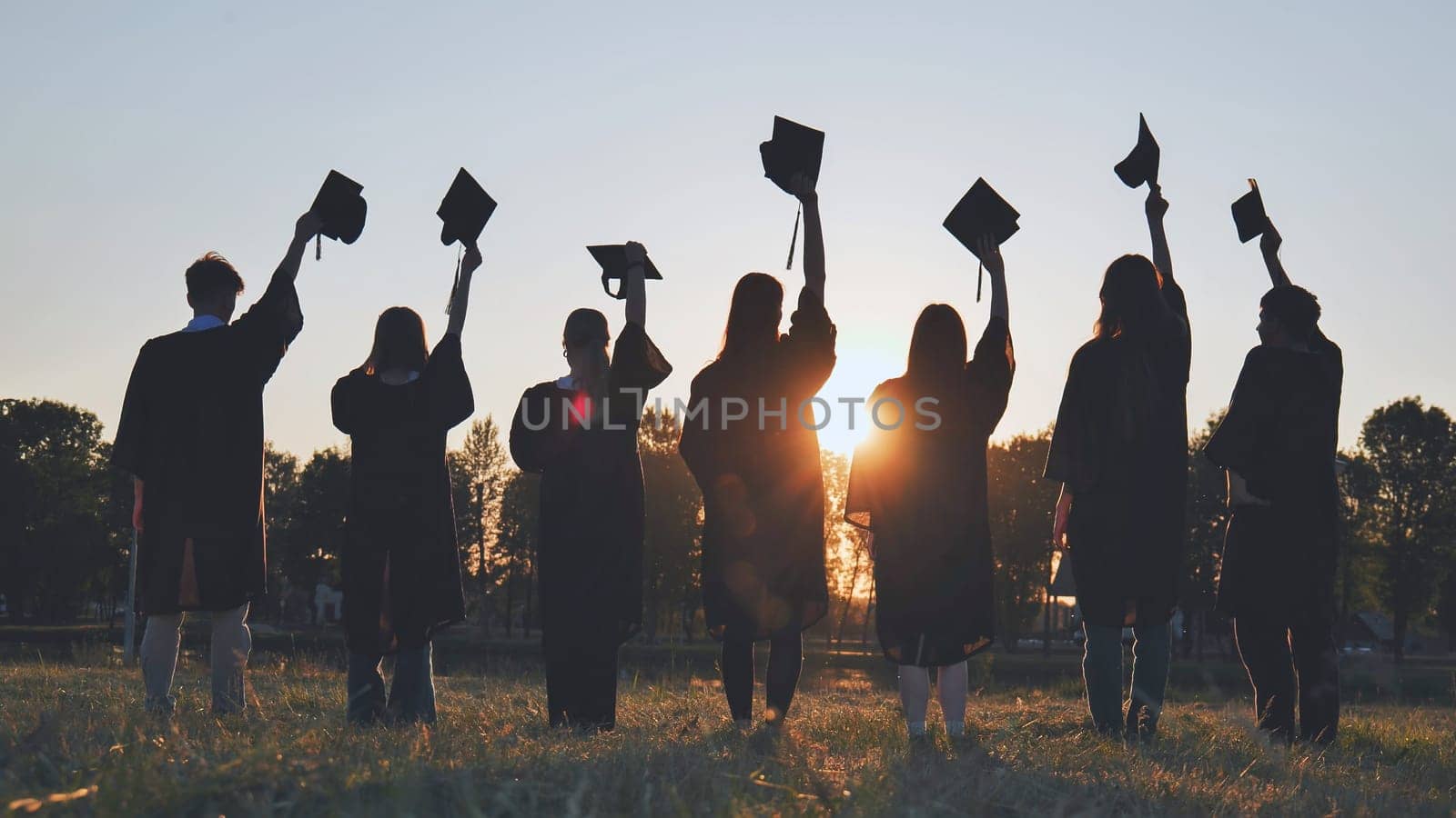 Silhouettes of college graduates waving their caps at sunset. by DovidPro