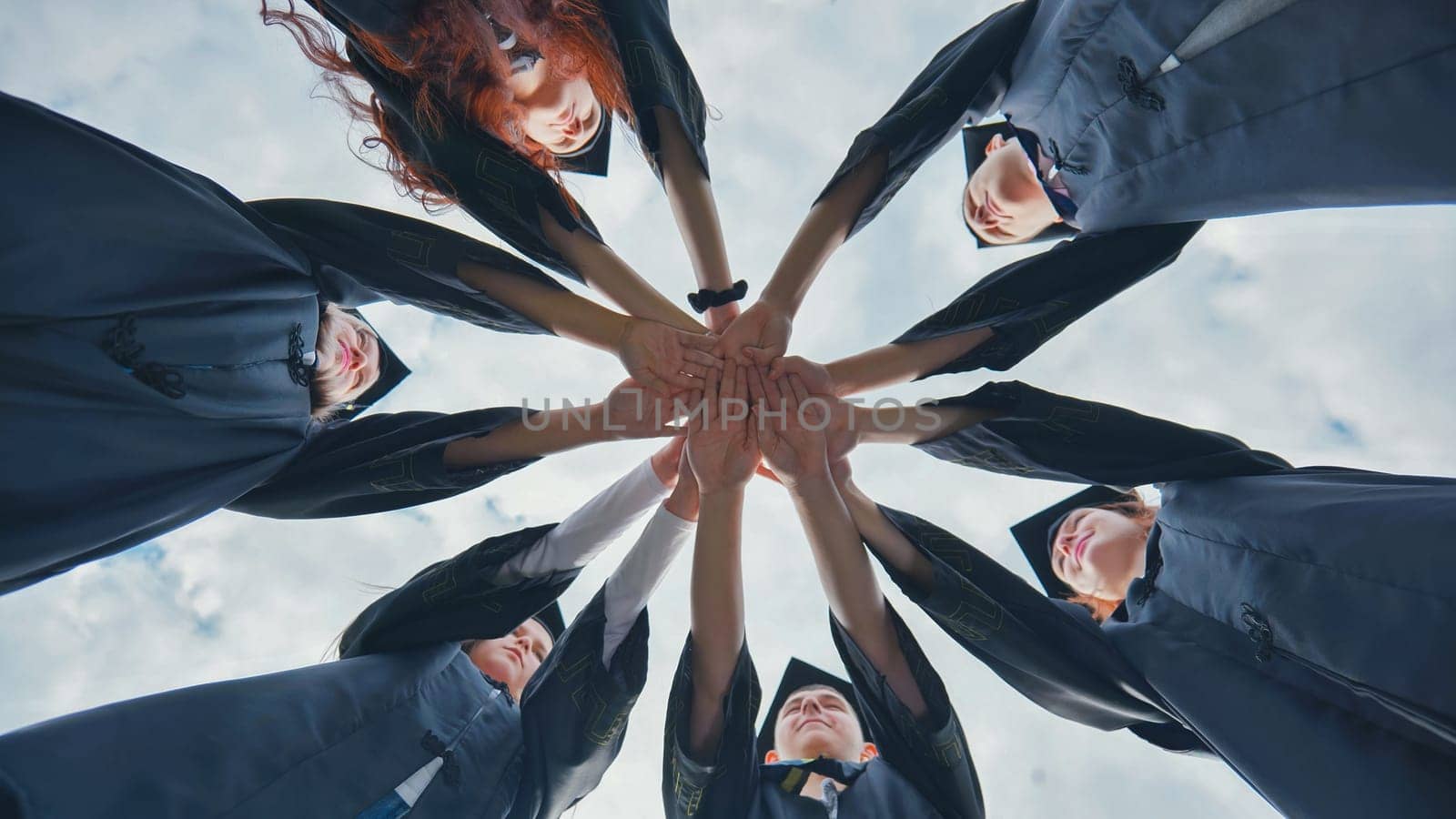 Team of college or university students celebrating graduation. Group of happy successful graduates in academic hats and robes standing in circle and putting their hands together.