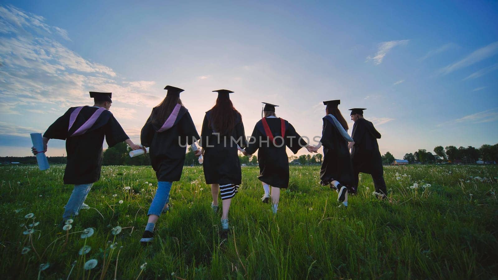 Six graduates in robes walk against the backdrop of the sunset