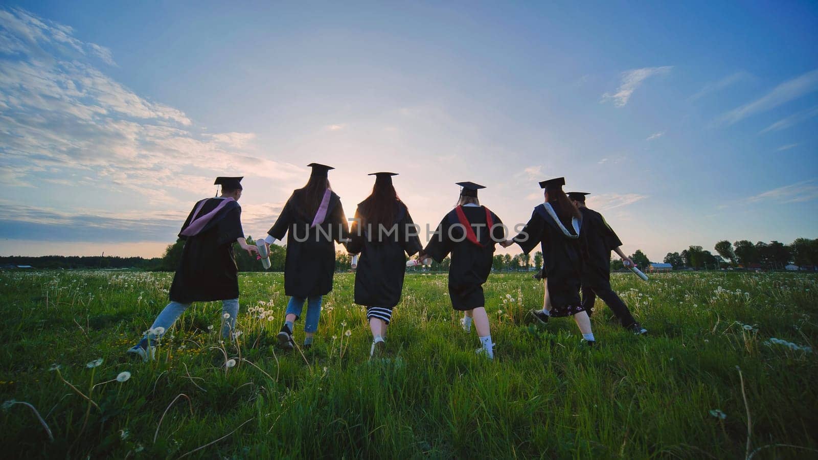 Six graduates in robes walk against the backdrop of the sunset