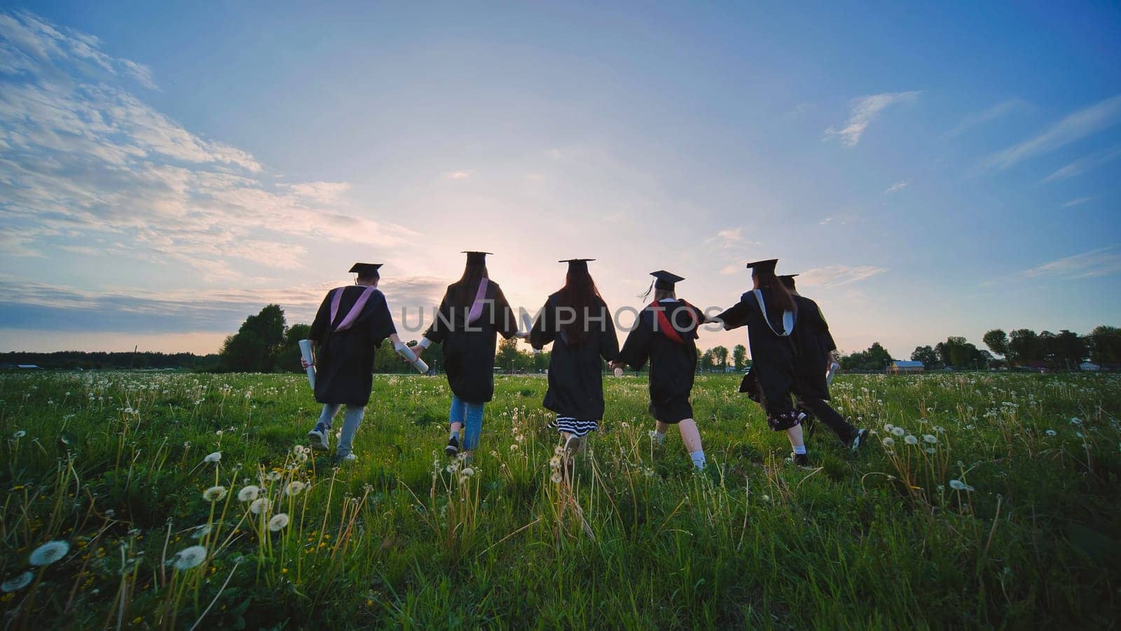 Six graduates in robes walk against the backdrop of the sunset