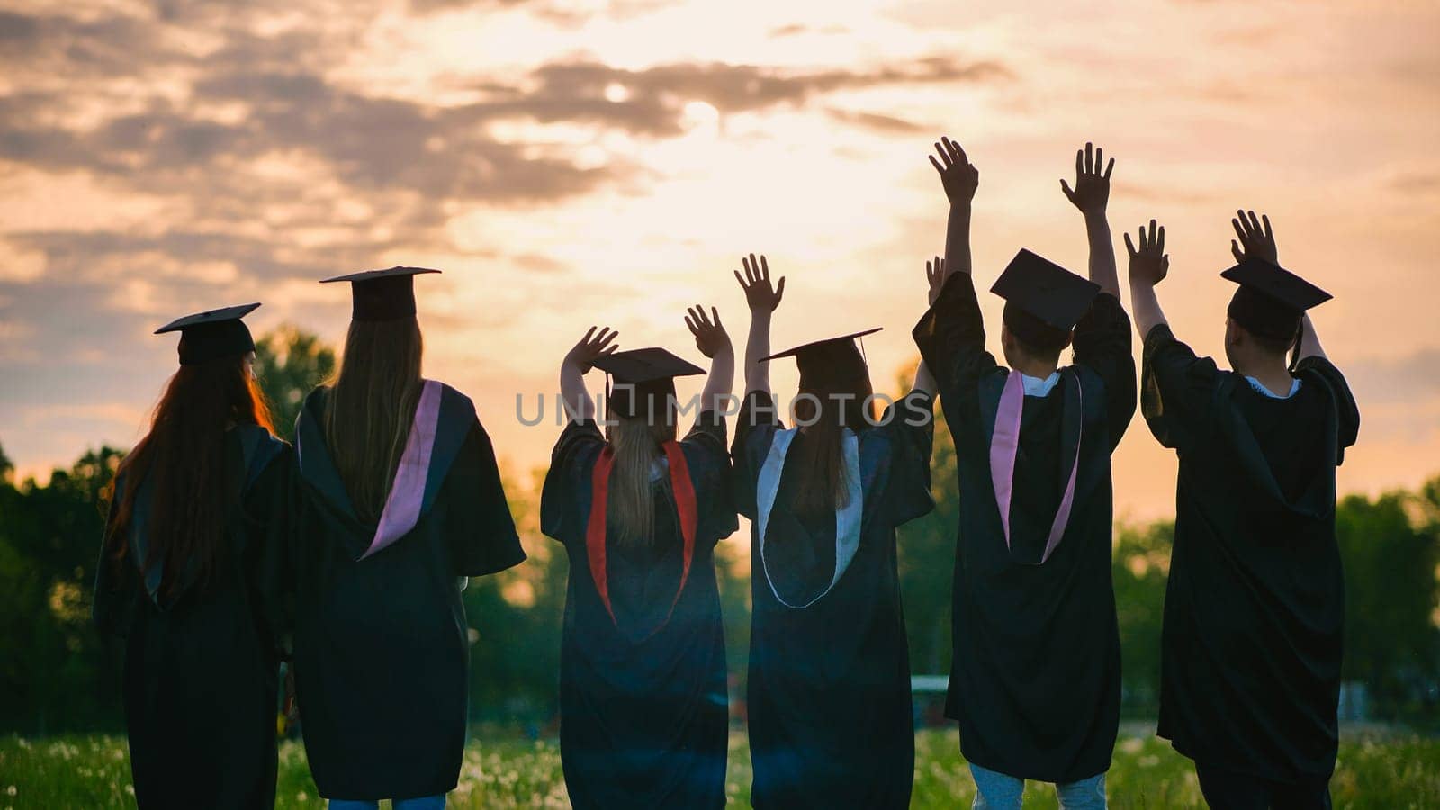 Silhouettes of graduates in black robes waving their arms against the evening sunset. by DovidPro