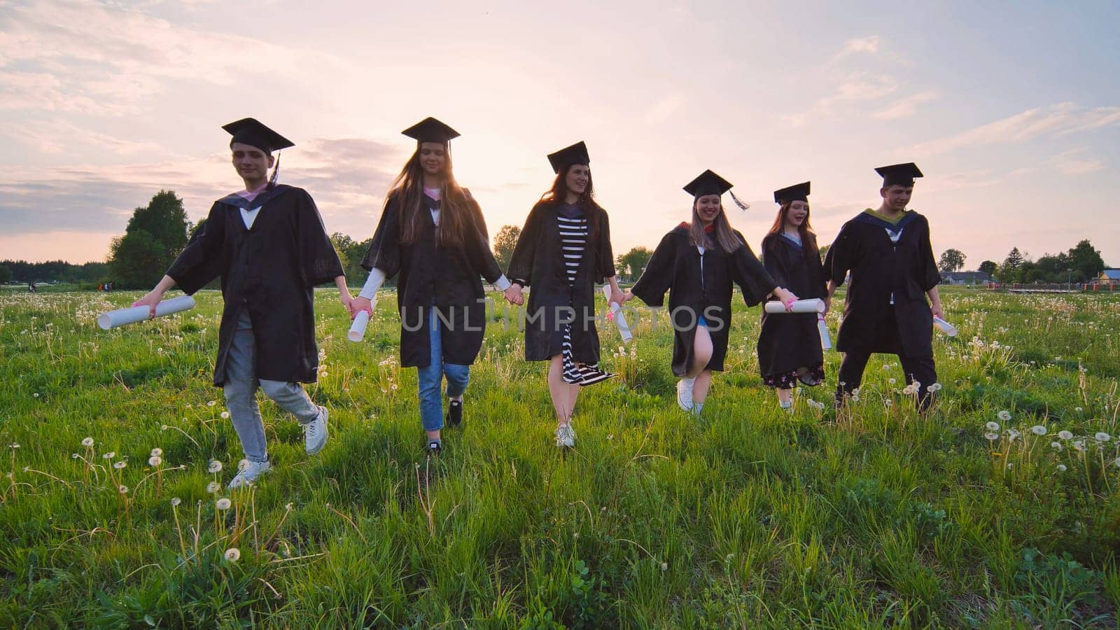 Six graduates in robes walk against the backdrop of the sunset. by DovidPro