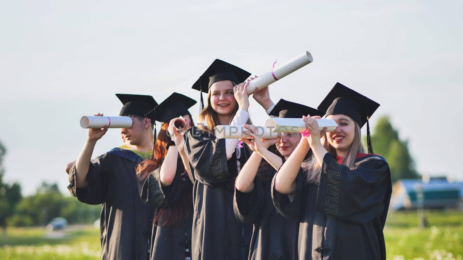 Cheerful graduates on a sunny day look through diplomas like a telescope. by DovidPro