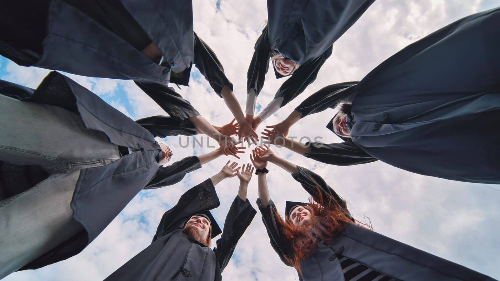 Group of happy successful graduates in academic hats and robes standing in circle and putting their hands together. Team of college or university students celebrating graduation. by DovidPro