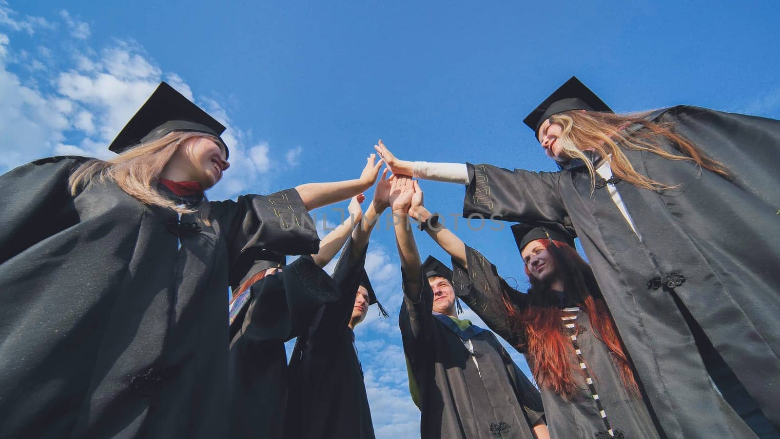 Group of happy successful graduates in academic hats and robes standing in circle and putting their hands together. Team of college or university students celebrating graduation. by DovidPro