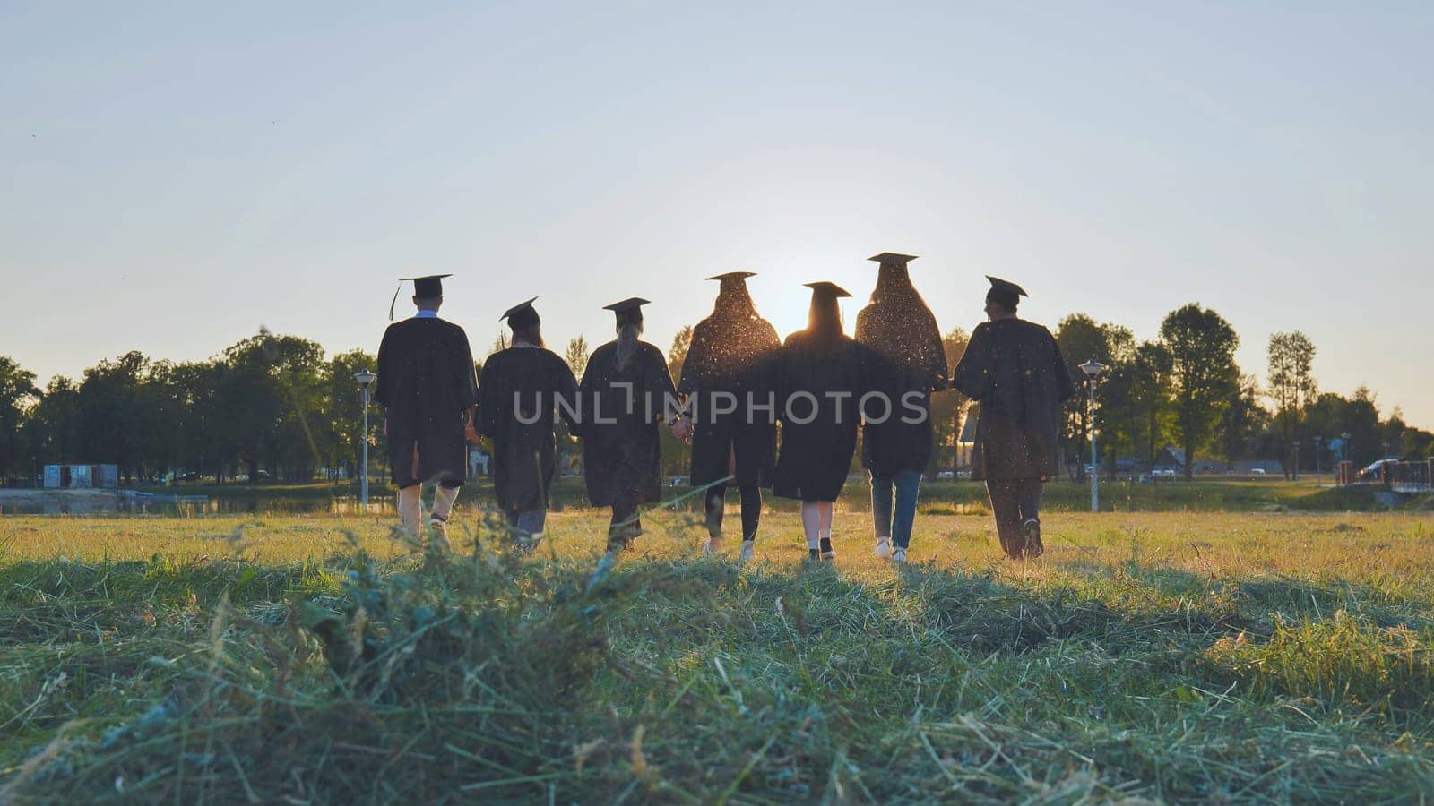 College graduates walk at sunset holding hands. by DovidPro