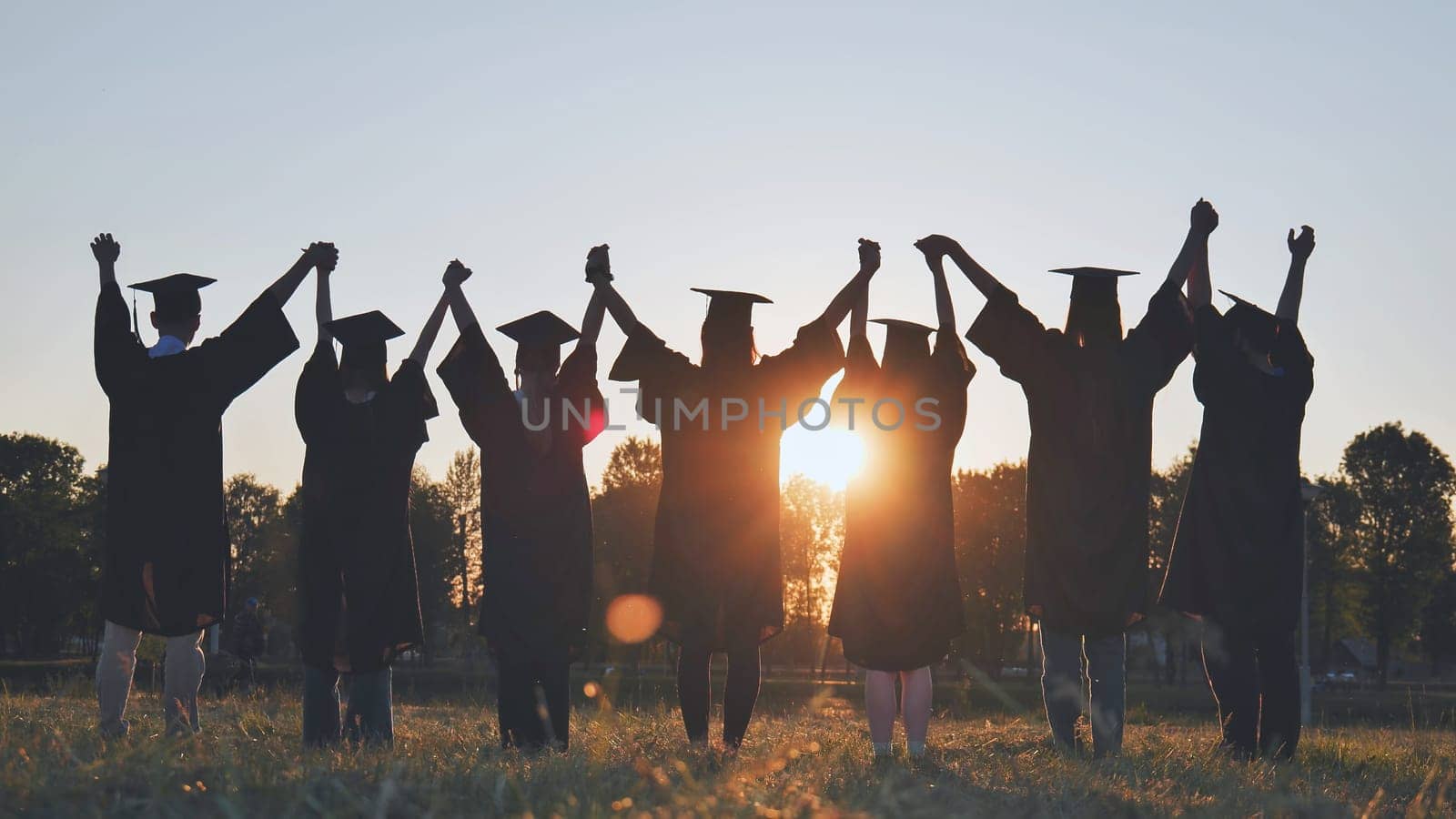 College graduates in robes holding hands at sunset. by DovidPro