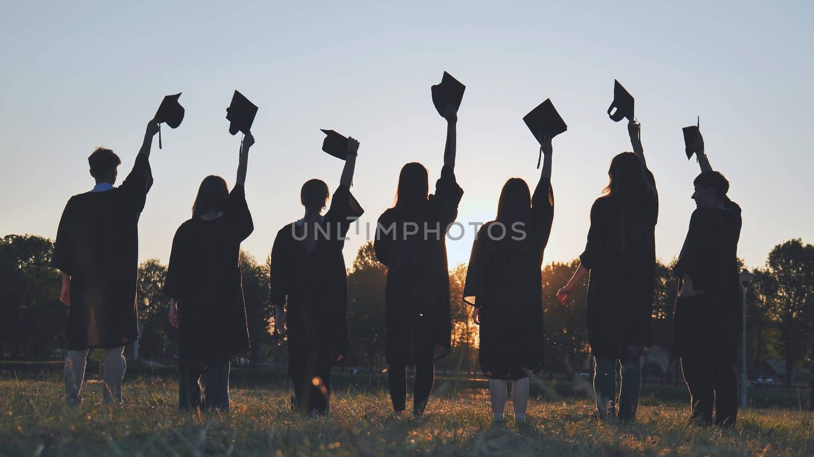 Silhouettes of college graduates waving their caps at sunset. by DovidPro