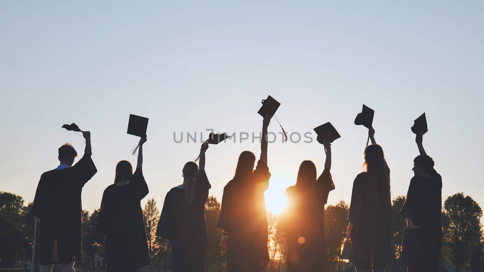 Silhouettes of college graduates waving their caps at sunset. by DovidPro