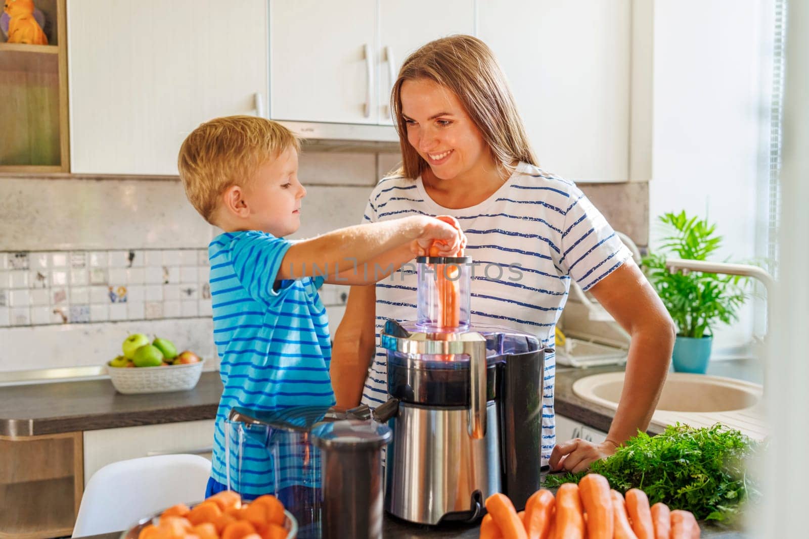 A young boy learns how to make carrot juice under his mother's guidance in a sunny home kitchen, surrounded by fresh ingredients.