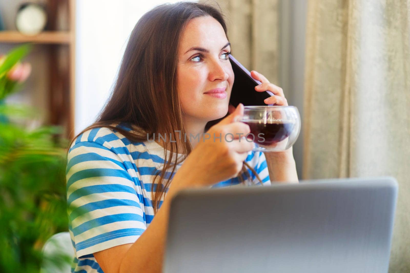 Thoughtful woman at home sipping coffee while having a conversation on her smartphone and working on her laptop.