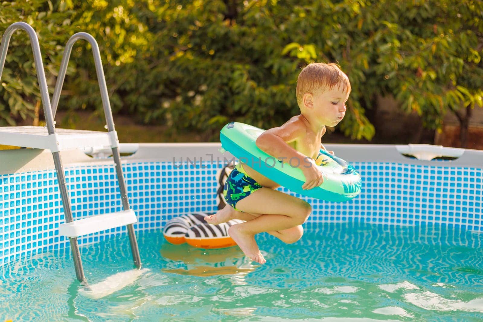 A little boy in swim trunks leaps into a swimming pool with a bright float ring on a sunny day, full of excitement.