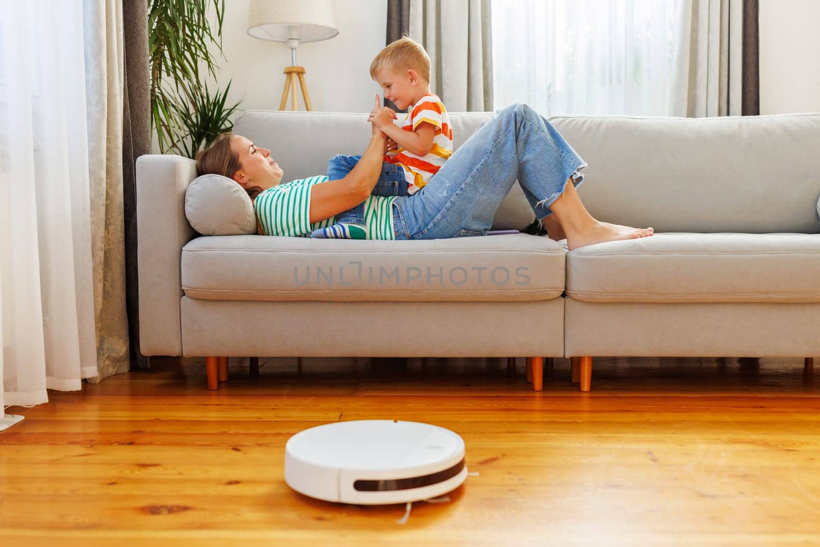 Joyful mother playing with her young son on a sofa in a clean, well-lit living room, while a robotic vacuum cleaner operates nearby