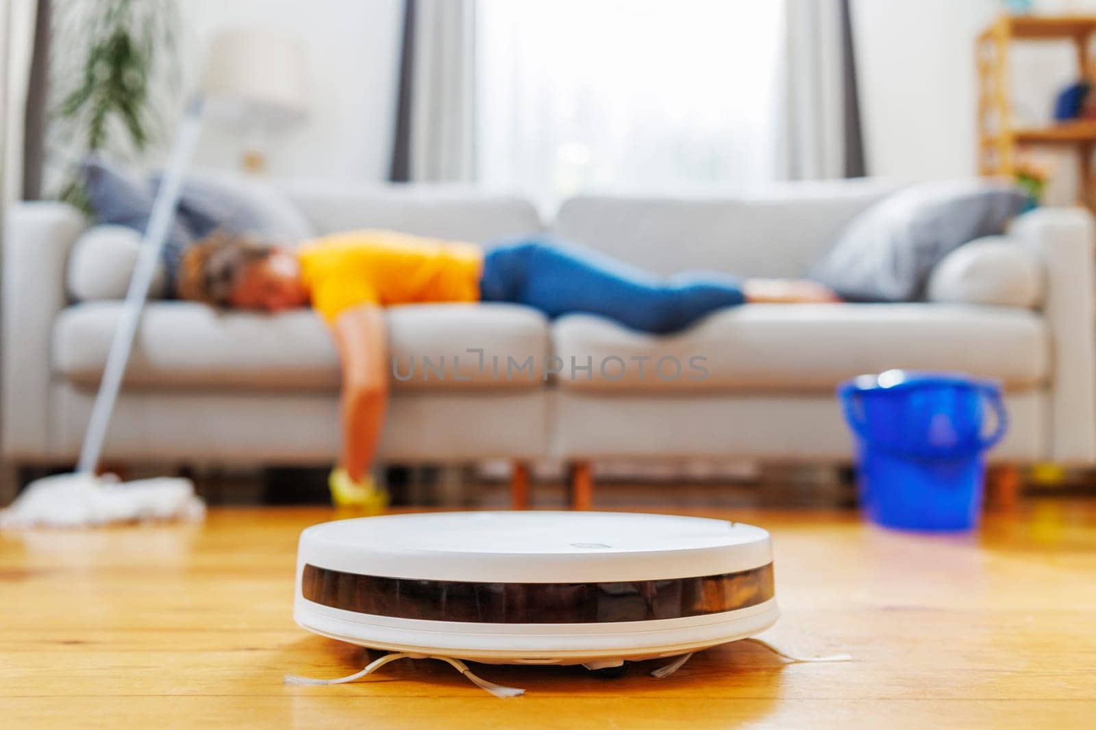 A robot vacuum cleaner is operating on a wooden floor in a living room where a woman is relaxing on the couch, symbolizing convenience and modern home cleaning technology.