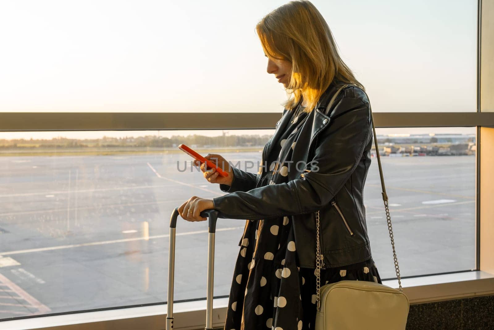 In the airport at sunset, a young woman, sporting a black jacket and polka-dot dress, is ready for boarding with her suitcase and checking her smartphone
