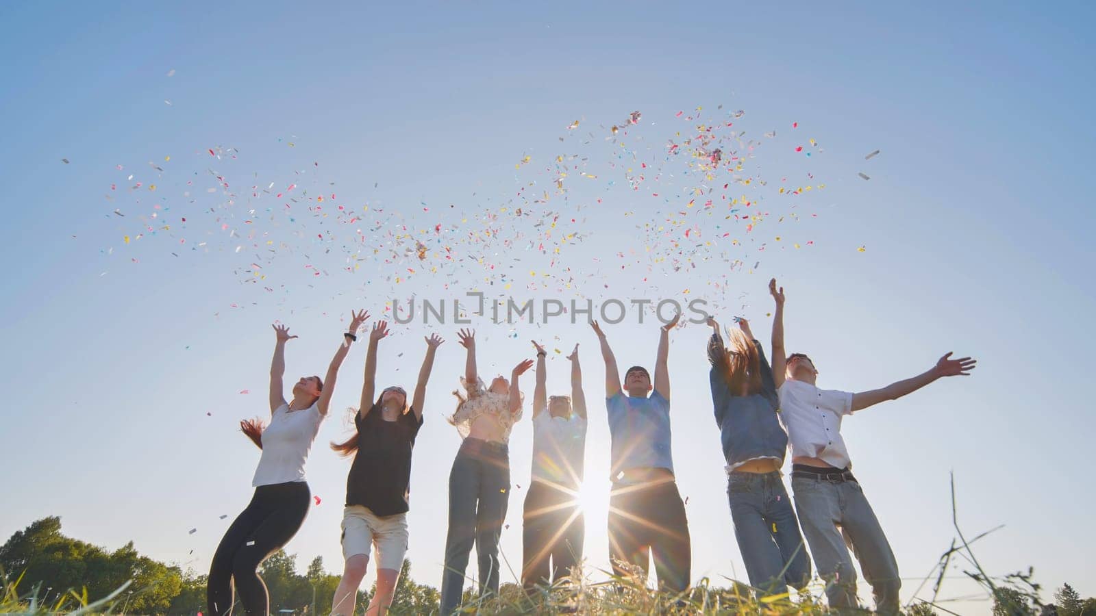 Friends toss colorful paper confetti from their hands against the rays of the evening sun. by DovidPro
