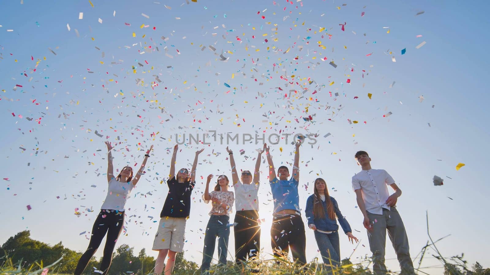 Friends toss colorful paper confetti from their hands against the rays of the evening sun
