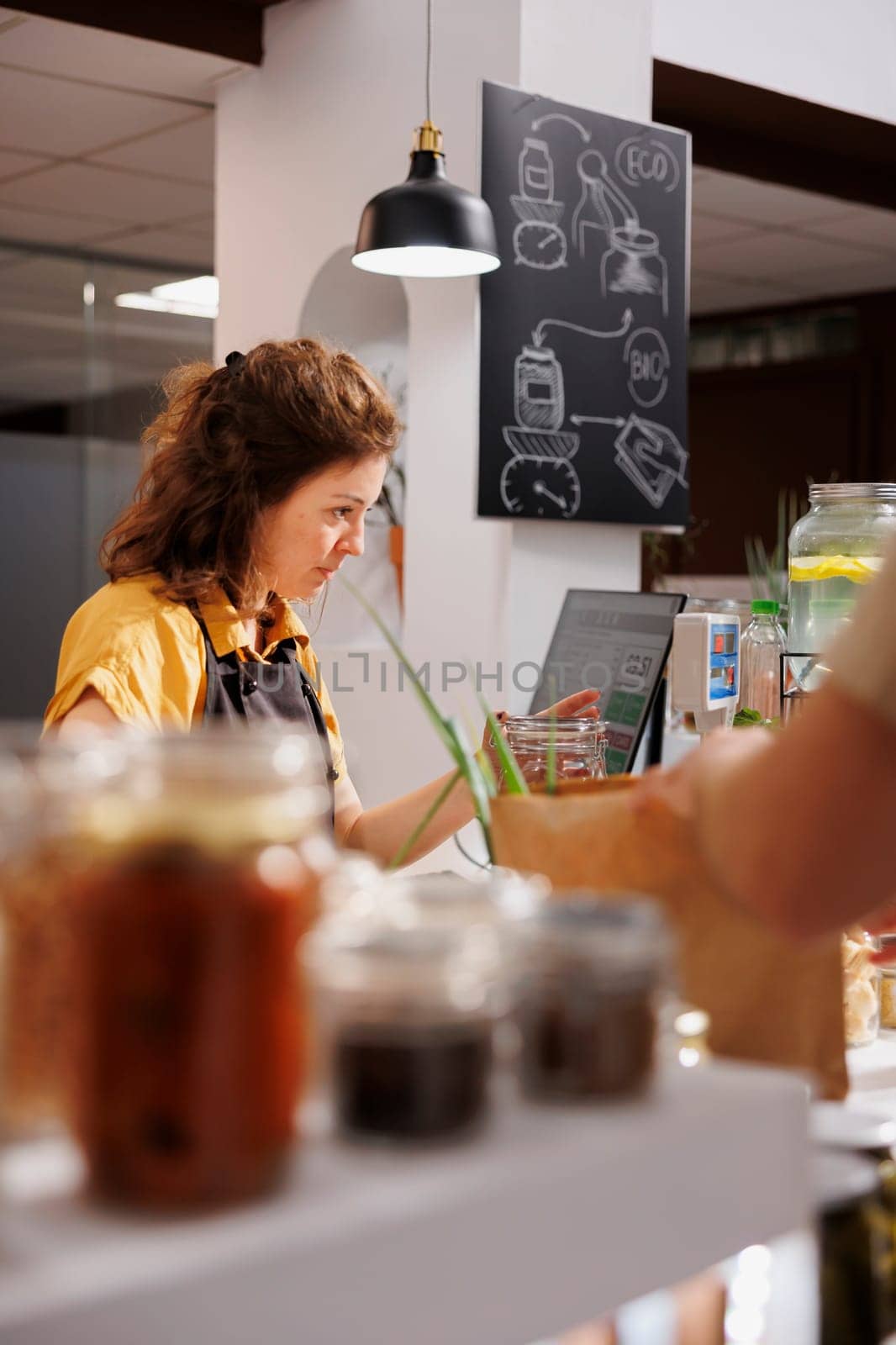 Storekeeper at zero waste supermarket checkout counter using bar code scanner to process customer transaction. Seller in sustainable local neighborhood shop scanning client food items