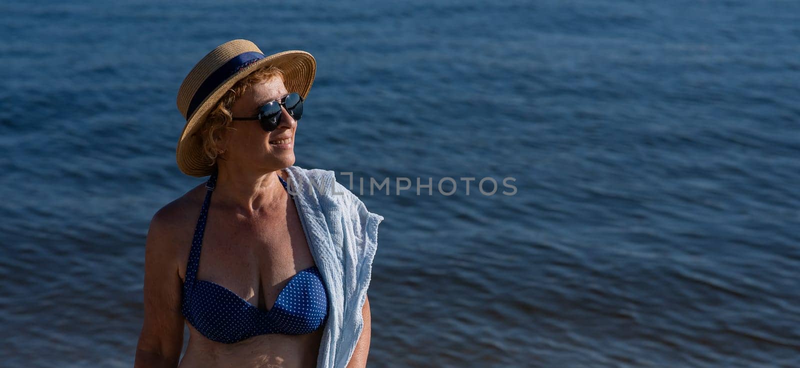 An old woman in a straw hat, sunglasses and a swimsuit is resting on the beach