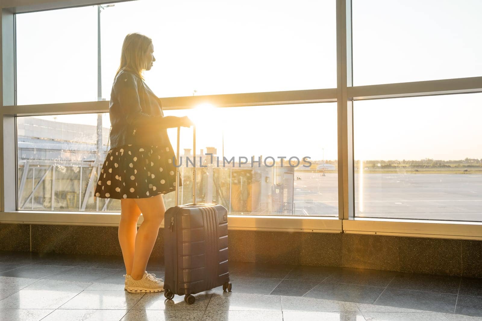 Young woman in a black jacket and polka-dot dress with a suitcase waiting for boarding in the airport at sunset.