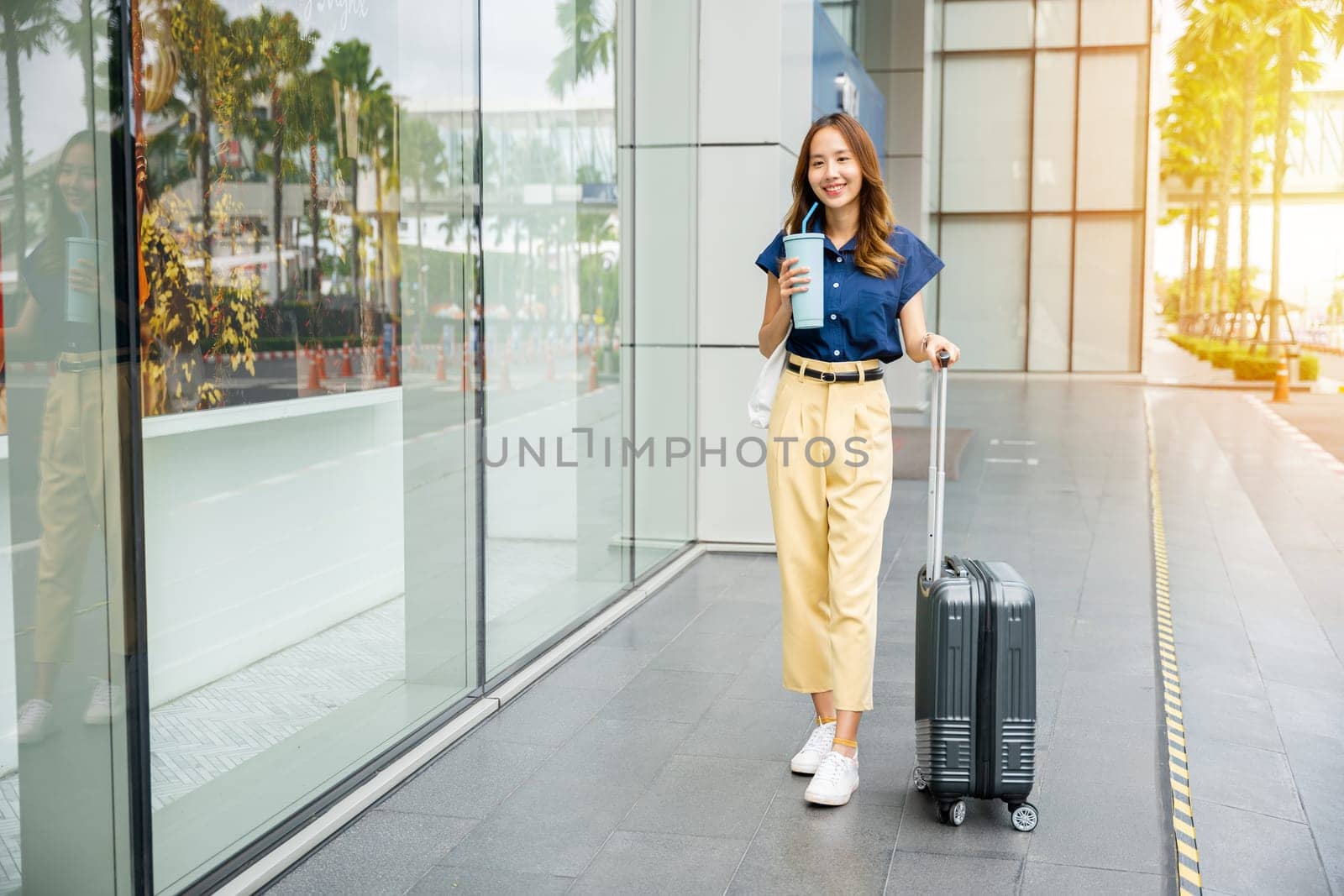 A young Asian woman with a sense of wanderlust holds a glass of water in one hand and her trusty suitcase in the other. Ready for travel, she sets off on her journey.