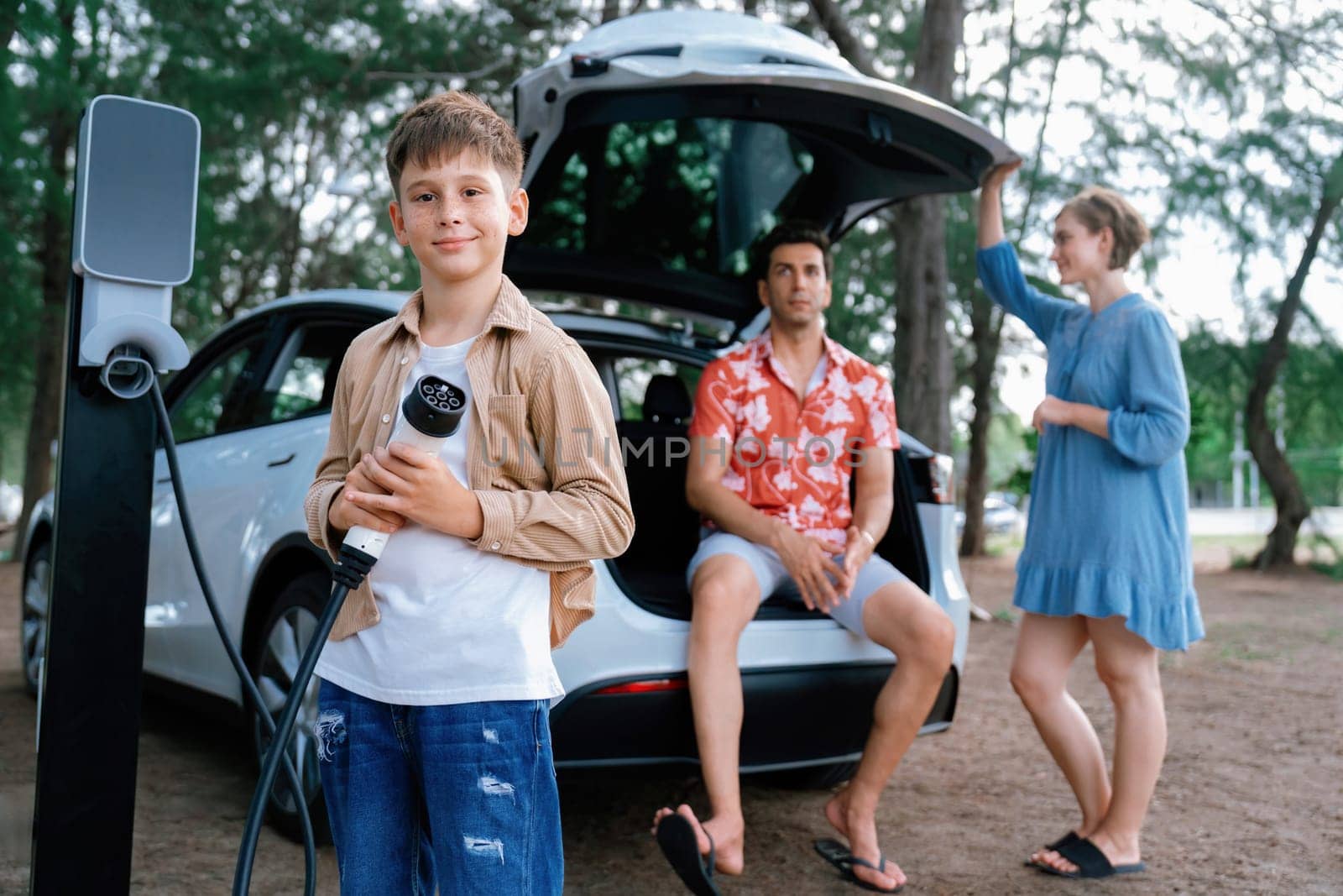 Little boy holding EV charger and point at camera with his family. Perpetual by biancoblue