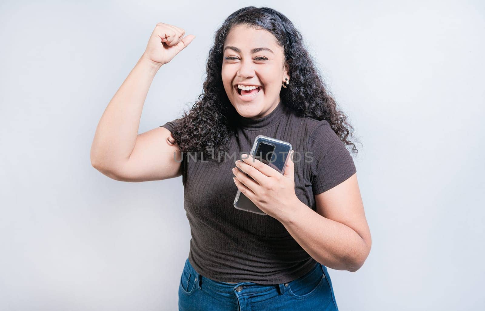 Winning happy young woman holding cell phone. Happy latin girl celebrating with phone isolated. Happy people holding smartphone and celebrating by isaiphoto