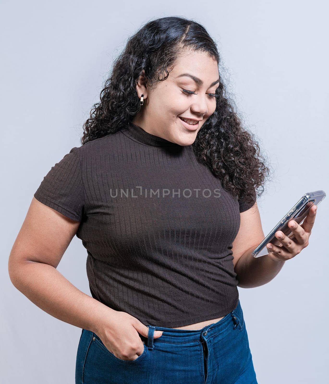 Smiling latin girl using cell phone isolated. Cheerful young woman with curly hair texting on cell phone isolated.