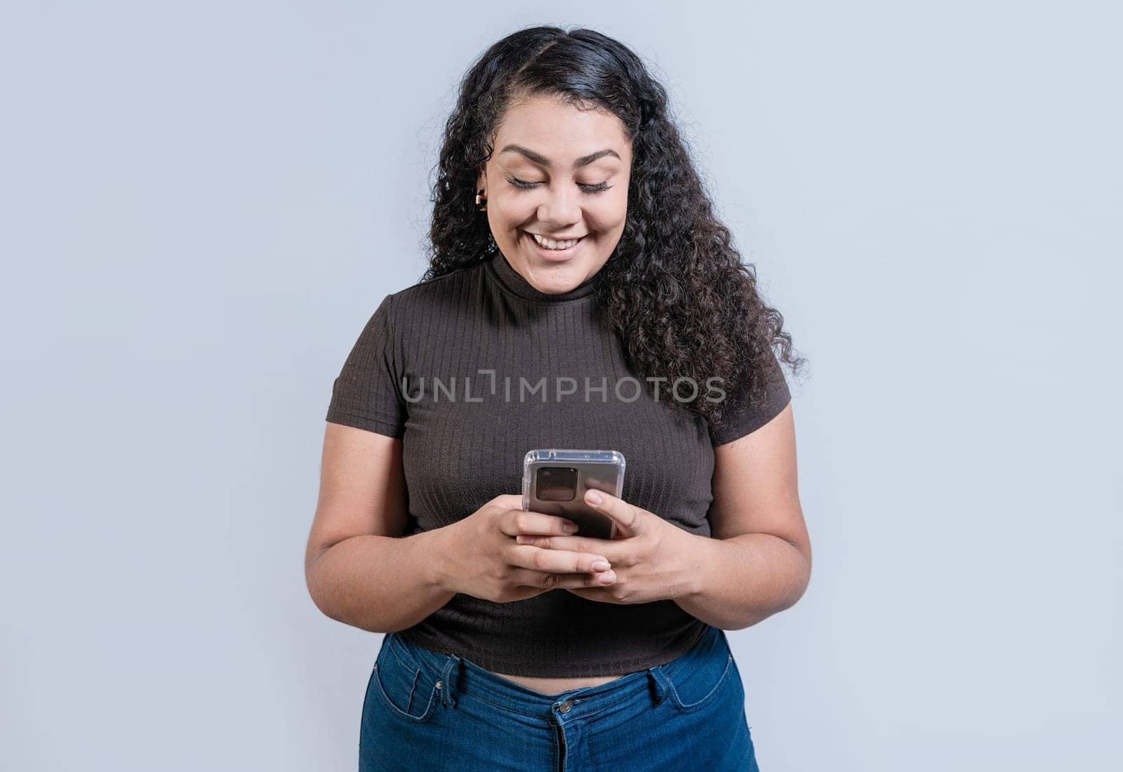 Cheerful young woman with curly hair texting on cell phone isolated. Smiling latin girl using cell phone isolated by isaiphoto