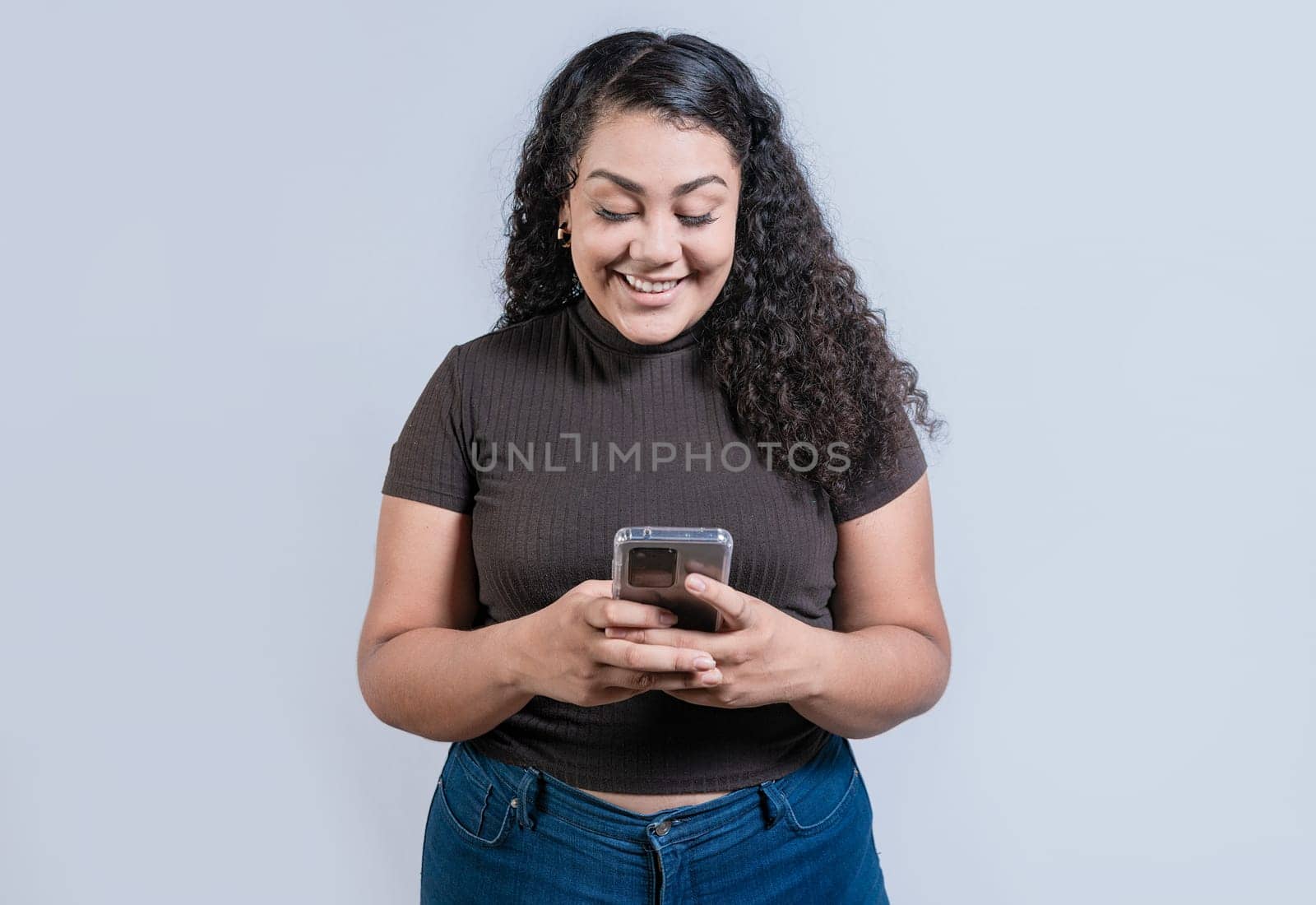 Cheerful young woman with curly hair texting on cell phone isolated. Smiling latin girl using cell phone isolated