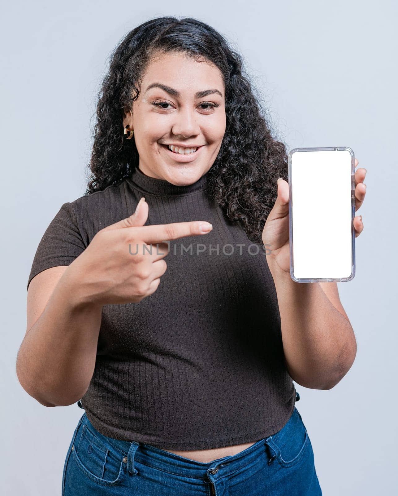 Smiling latin girl showing and pointing at cell phone screen isolated. Funny girl with curly hair showing an advertisement on her cell phone by isaiphoto