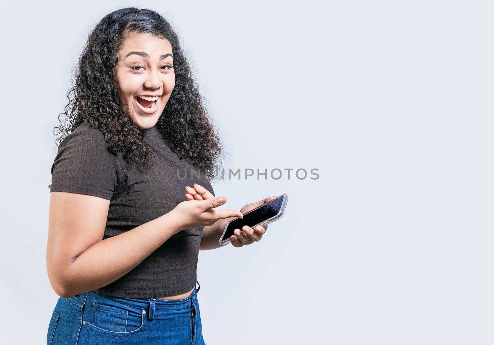 Smiling young woman holding cell phone and looking at camera. Happy latin girl using and pointing cell phone looking at camera isolated by isaiphoto