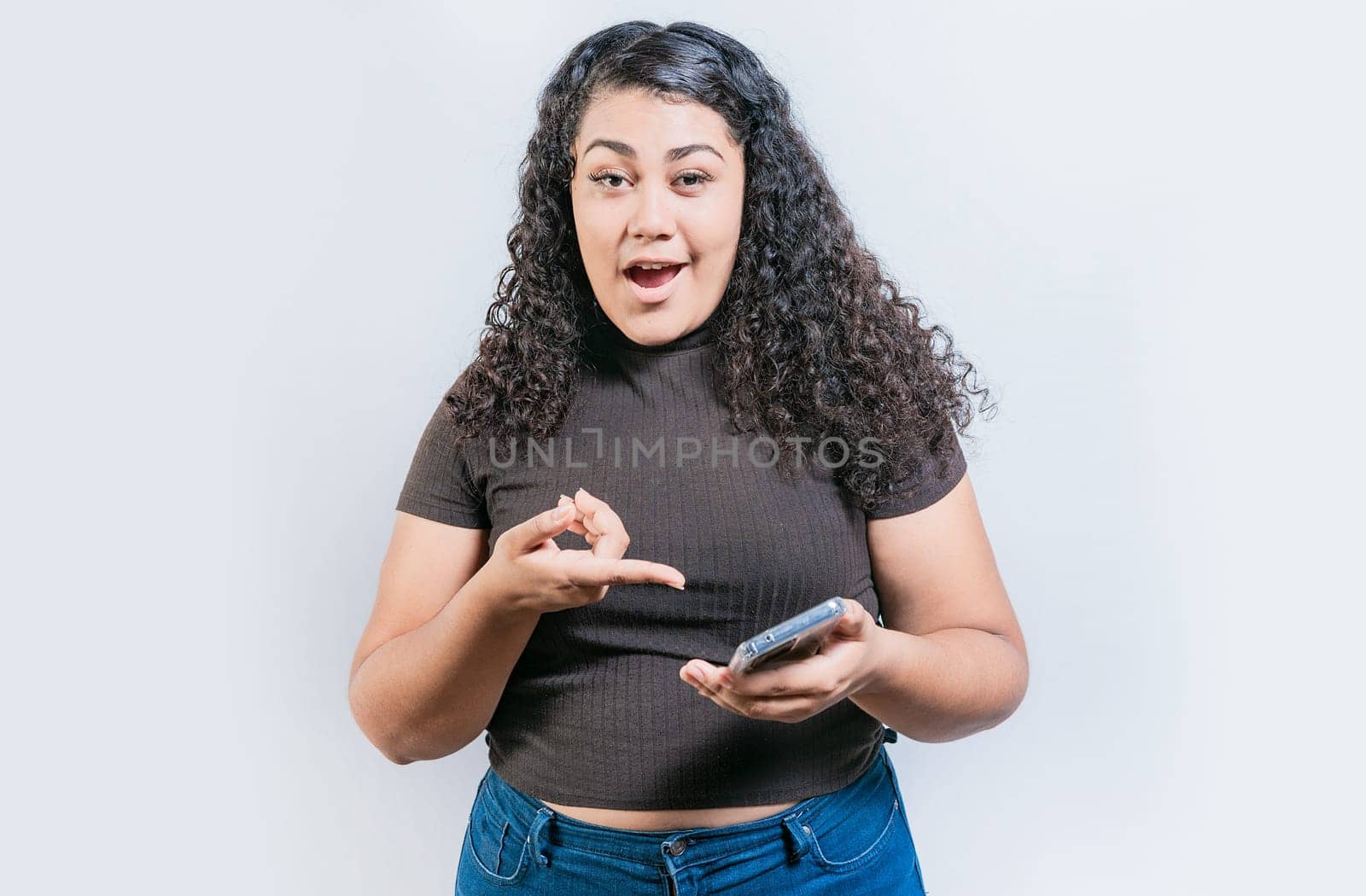 Amazed girl using and pointing cell phone looking at camera isolated. Portrait of surprised latin woman holding cell phone and looking at camera by isaiphoto