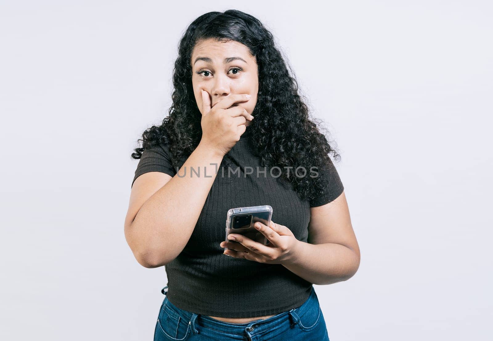 Surprised young woman covering mouth holding phone isolated. Astonished young girl holding phone and covering mouth isolated by isaiphoto
