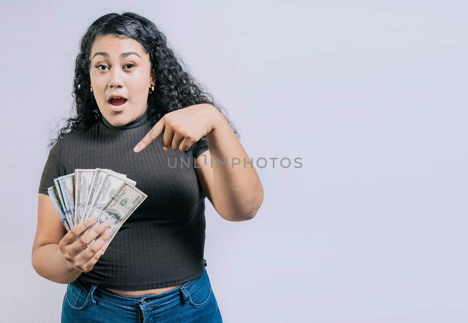 Amazed young woman holding and pointing dollars banknotes isolated. Surprised latin girl holding dollar cash isolated