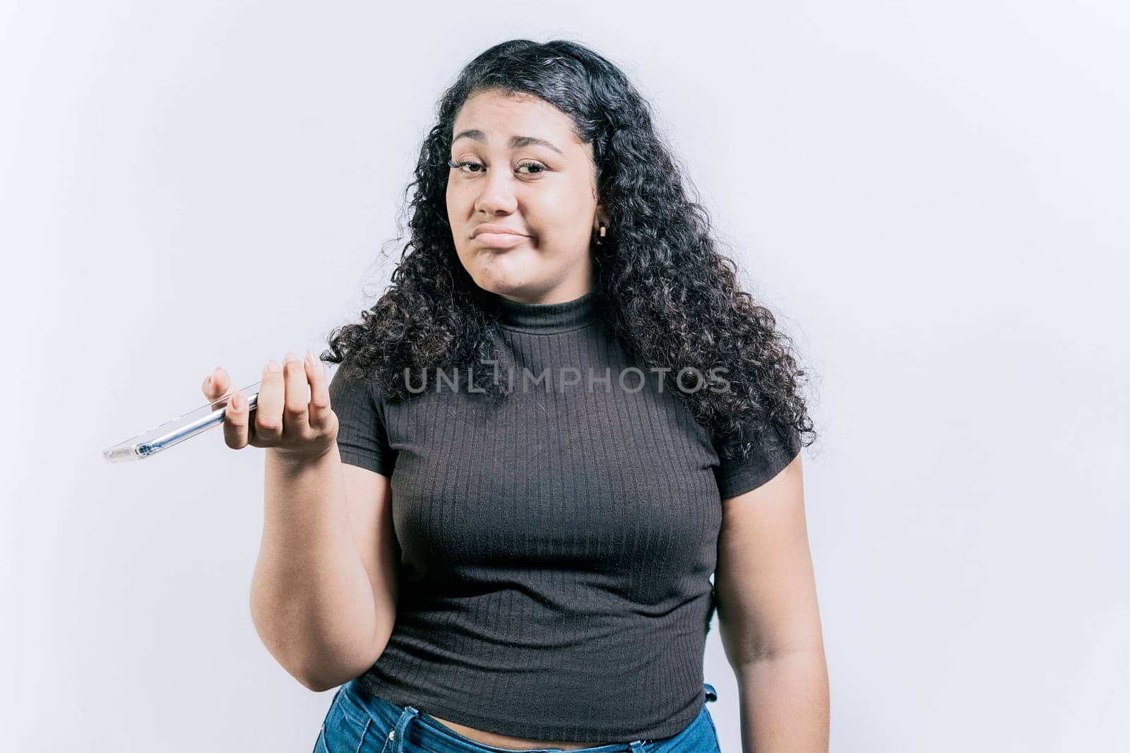 Indifferent young woman holding cell phone isolated. Sad woman with cell phone looking at camera. Young skeptical woman with cell phone isolated by isaiphoto