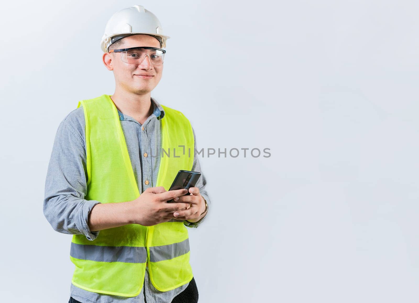 Smiling engineer holding phone looking at camera. Latin male engineer using phone isolated. Young engineer in vest and helmet using cell phone isolated by isaiphoto