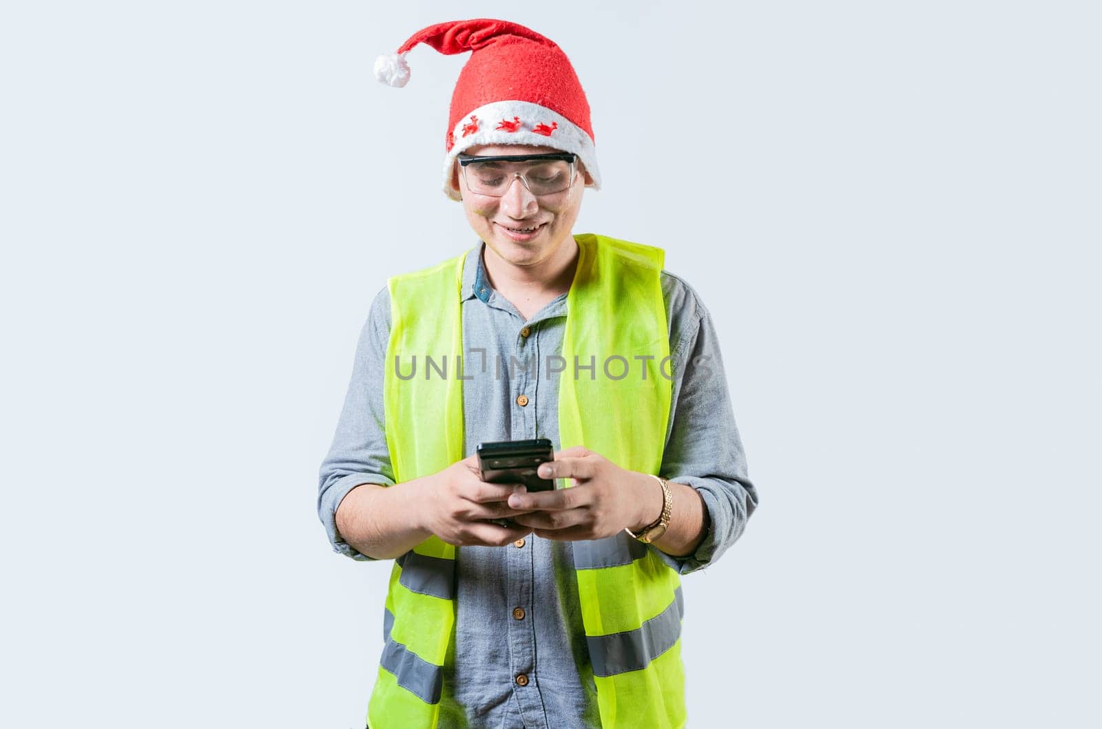 Smiling engineer in christmas hat using cell phone isolated. Cheerful young engineer in christmas hat using smartphone