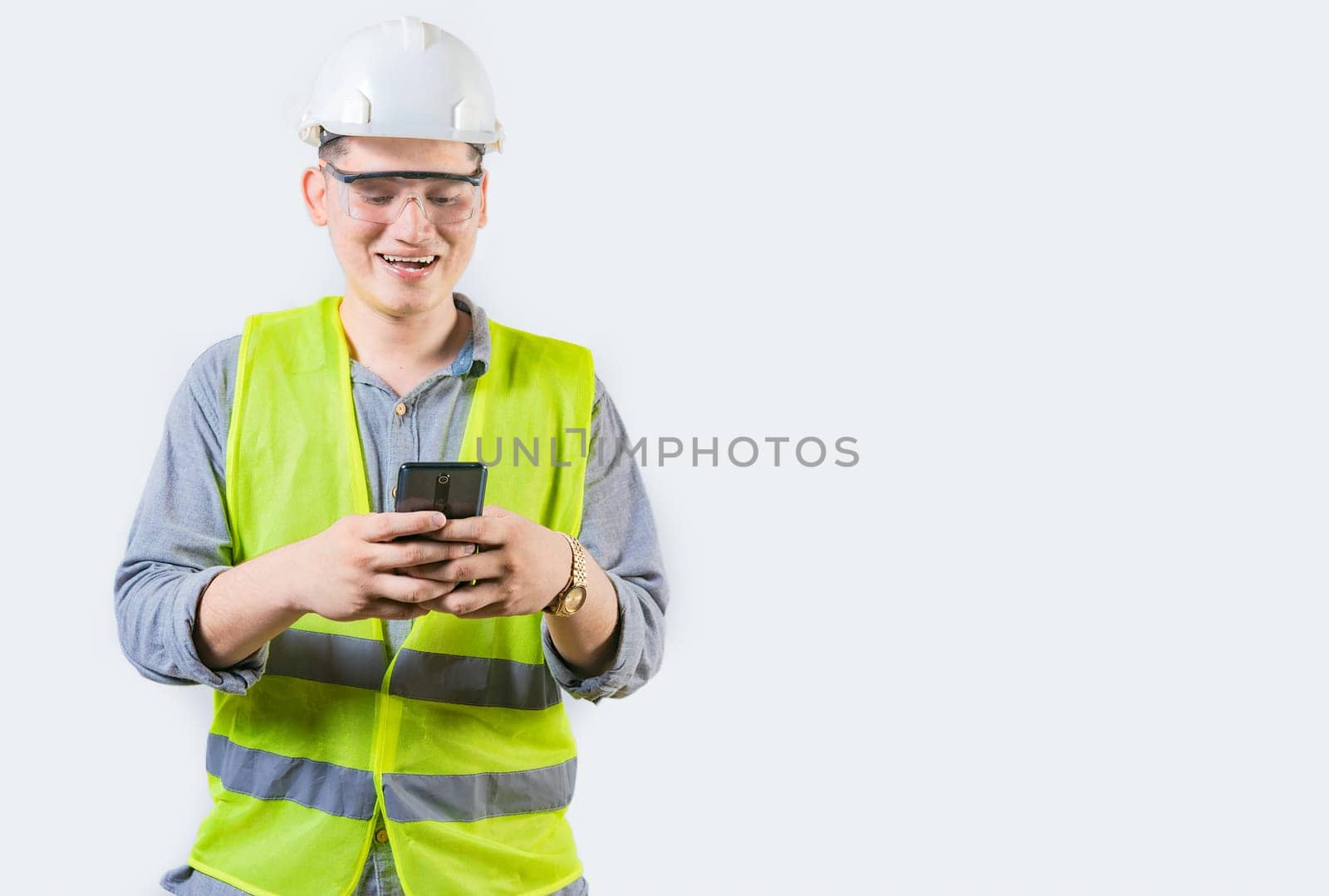 Handsome engineer holding phone isolated. Cheerful builder engineer texting phone on isolated background. Young engineer in vest and helmet using cell phone