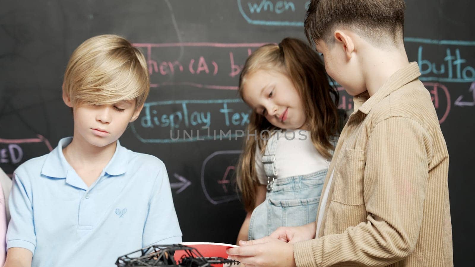 Group of caucasian student checks robotic model while taking a note.at blackboard with engineering code or prompt written. Children studying, learning about programing robot in STEM class. Erudition.
