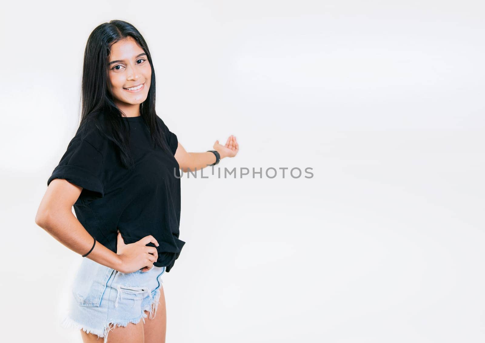 Happy young girl welcoming you isolated. Smiling latin girl pointing back presenting a product. Latin girl pointing a promotion with her palm by isaiphoto