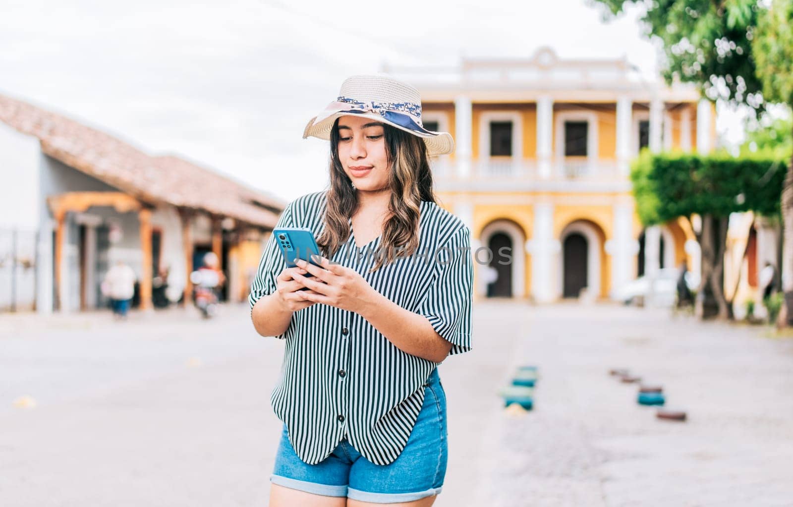 Beautiful young tourist woman in hat using phone on the street. Latin tourist girl texting with phone on the street by isaiphoto