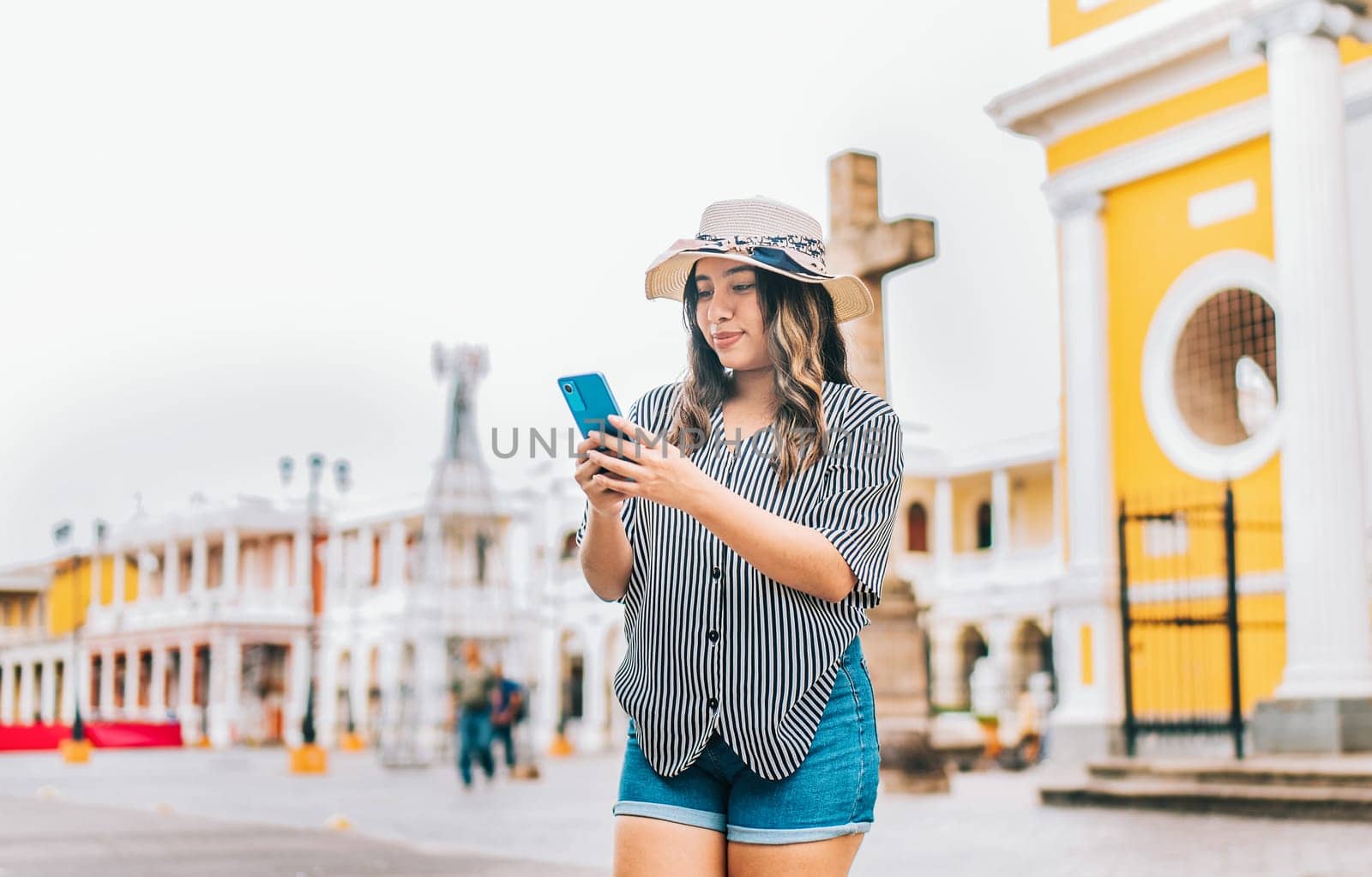 Female tourist texting phone in the square of Granada. Smiling young tourist in hat using cell phone on the street by isaiphoto