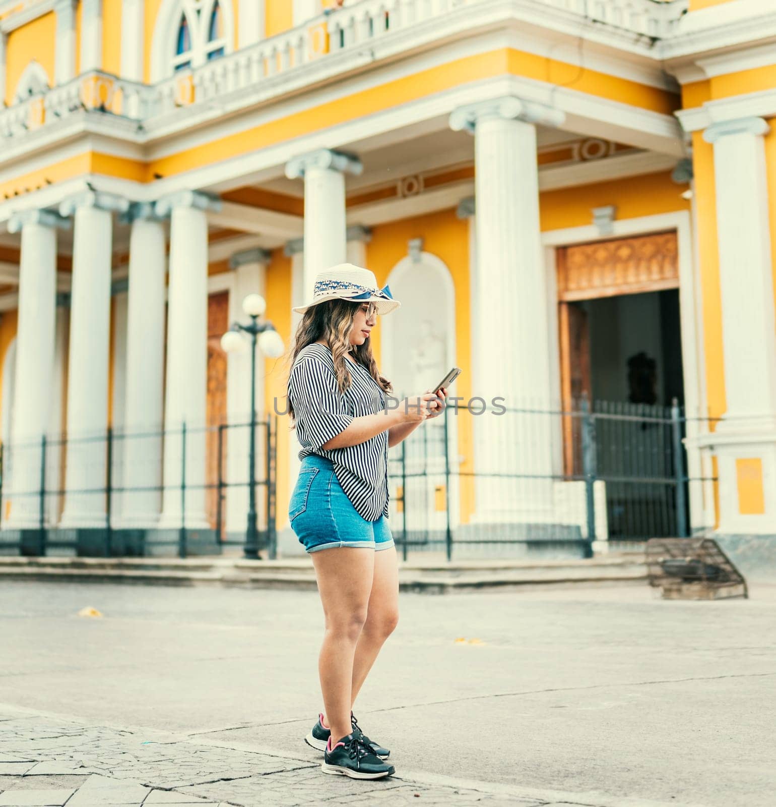 Lifestyle of tourist girl texting with phone on the street of square. Beautiful young travel girl with hat using cell phone on the street of Granada Nicaragua by isaiphoto