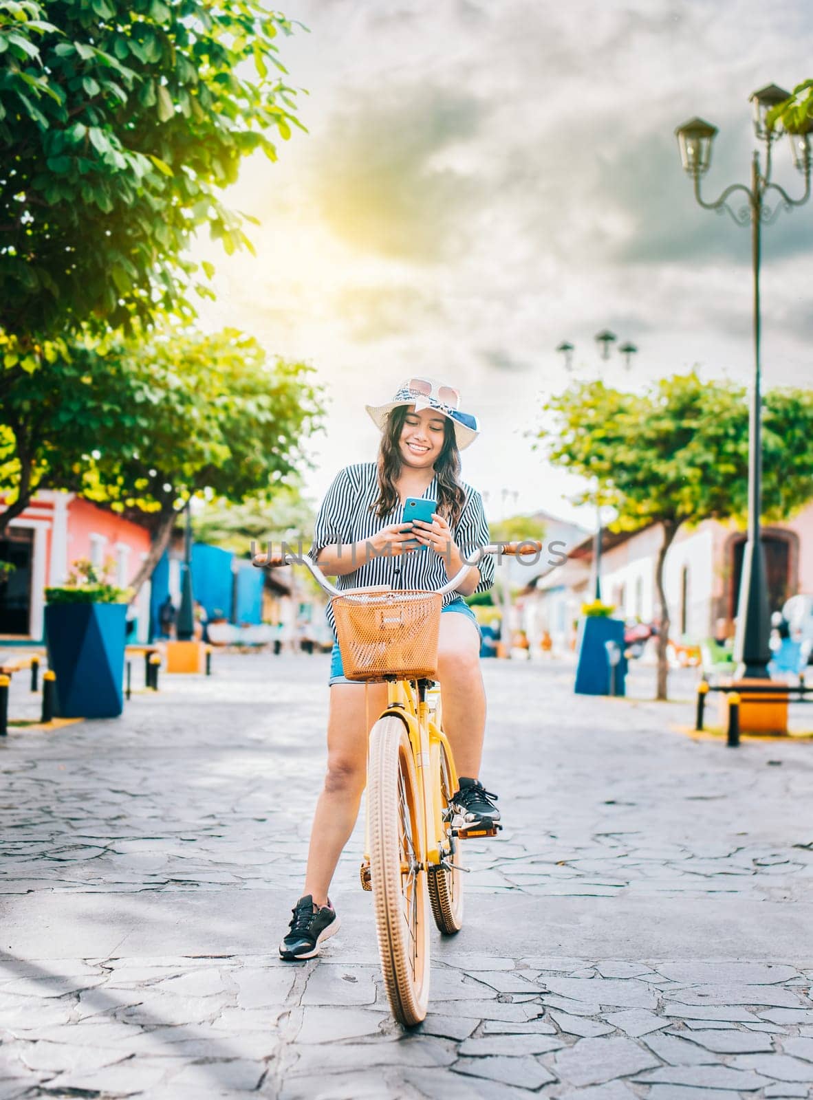 Happy tourist girl on bicycle using cell phone on the street. Beautiful girl in hat on bicycle with cell phone on the street of La Calzada, Granada, Nicaragua by isaiphoto